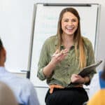 A smiling young female creative professional sits on a stool before a group of employees.  She gestures as she speaks to a new employee training class.